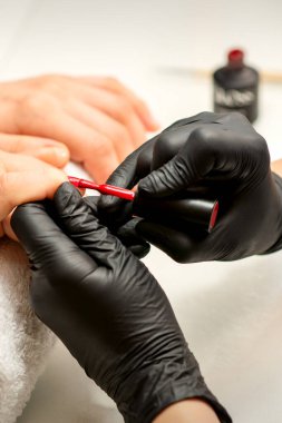 Manikür cila resmi. Kauçuk siyah eldivenli bir manikür ustasının yakın çekimi güzellik salonundaki bir kadının tırnağına kırmızı cila sürüyor.