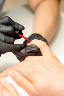 Manikür cila resmi. Kauçuk siyah eldivenli bir manikür ustasının yakın çekimi güzellik salonundaki bir kadının tırnağına kırmızı cila sürüyor.