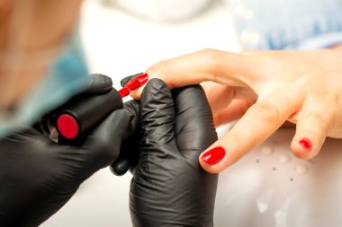 Manikür cila resmi. Kauçuk siyah eldivenli bir manikür ustasının yakın çekimi güzellik salonundaki bir kadının tırnağına kırmızı cila sürüyor.