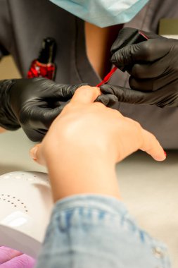 Manikür cila resmi. Kauçuk siyah eldivenli bir manikür ustasının yakın çekimi güzellik salonundaki bir kadının tırnağına kırmızı cila sürüyor.