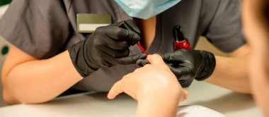 Manikür cila resmi. Kauçuk siyah eldivenli bir manikür ustasının yakın çekimi güzellik salonundaki bir kadının tırnağına kırmızı cila sürüyor.