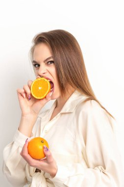 Genç, beyaz, kurnaz esmer kadın turuncu yarısını ısırıyor ve beyaz arka planda beyaz bir gömlek giymiş kameraya bakıyor.