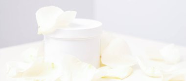 White jar with cream among delicate white rose flowers petals on a light background. Natural organic cosmetics concept, close up, mock-up