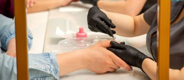 Manikür ustası, manikür salonunda koruyucu eldiven takan kadın tırnaklarından makas çıkartıyor.