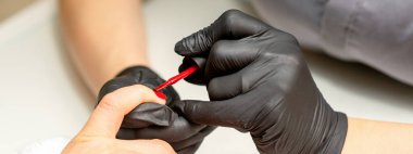 Manikür cila resmi. Kauçuk siyah eldivenli bir manikür ustasının yakın çekimi güzellik salonundaki bir kadının tırnağına kırmızı cila sürüyor.