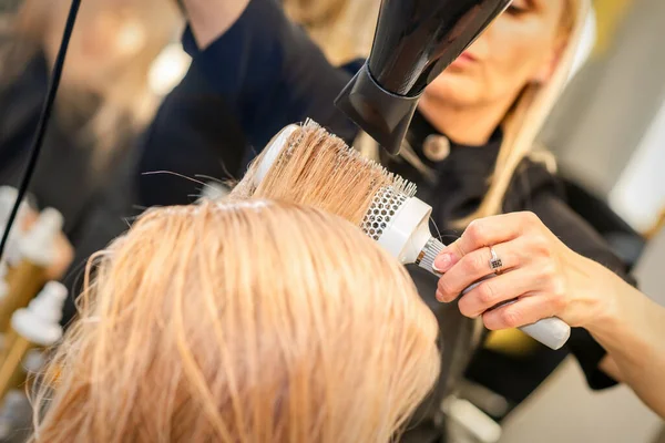 Secagem de cabelo no estúdio de cabeleireiro. estilista de cabeleireira  seca o cabelo com secador de cabelo e escova redonda ruiva de uma mulher em  um salão de beleza.