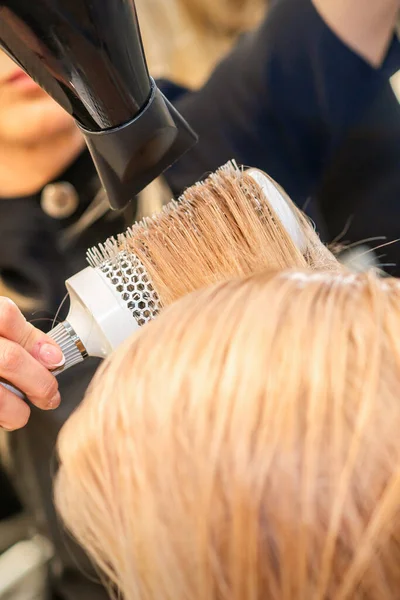 Secagem de cabelo no estúdio de cabeleireiro. estilista de cabeleireira  seca o cabelo com secador de cabelo e escova redonda ruiva de uma mulher em  um salão de beleza.