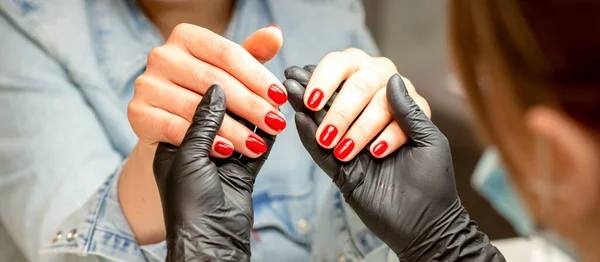 マニキュア職人はネイルサロンで完成した赤いポリッシュマニキュアを示す美しい若い女性の手を保持しています — ストック写真
