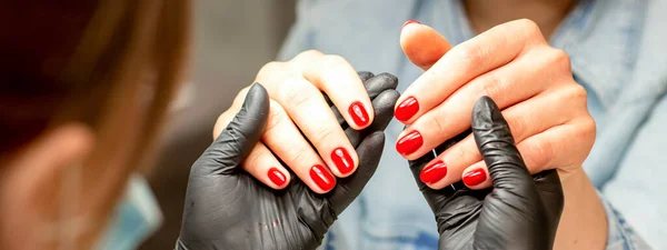 マニキュア職人はネイルサロンで完成した赤いポリッシュマニキュアを示す美しい若い女性の手を保持しています — ストック写真