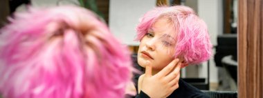 Yeni kısa pembe saç stili olan güzel beyaz kadın aynadaki yansımasına bakıyor kuaför salonundaki saç stilini kontrol ediyor.