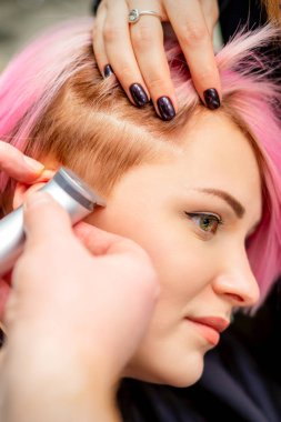 Hairdresser shaves female temple with pink hair by electric shaver in a hairdresser salon, close up