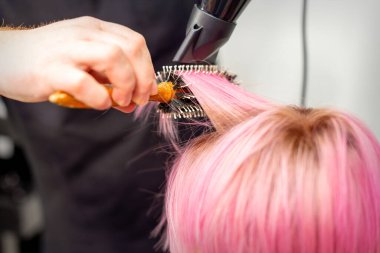 Siyah saç kurutma makinesi ve siyah yuvarlak fırçası olan beyaz bir kadının kısa pembe saçlarını bir kuaförde erkek bir kuaförün elleriyle kurutmak.