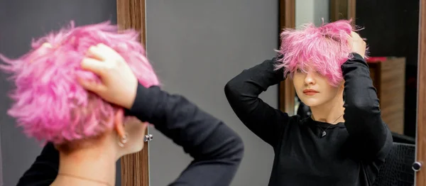 年轻女子在理发店的镜子前 用手检查着她新的卷曲的短粉色发式 — 图库照片
