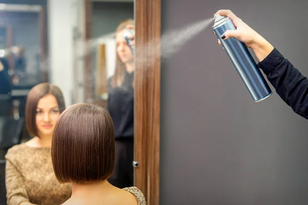 A hairdresser is using hair spray to fix the short hairstyle of the young brunette woman sitting in the hair salon