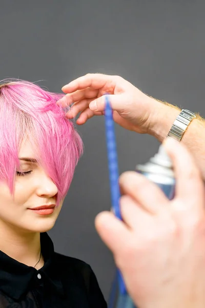 The hairdresser is using hair spray to fix the short pink hairstyle of the young caucasian woman in the hair salon