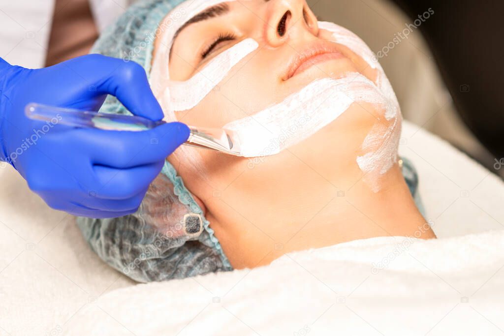 The beautician with brush applies a photochemical and glycolic peeling face mask to the female patient face in the beauty clinic