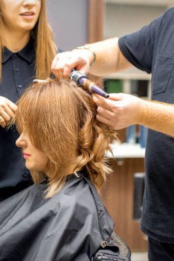 İki kuaför, kuaförde saç maşasıyla uzun kahverengi saç stili yapar.