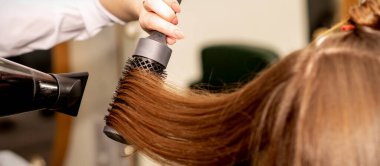 Bir kuaför, uzun kahverengi saçları bir saç kurutma makinesi ve bir güzellik salonunda yuvarlak fırçayla kurutuyor.