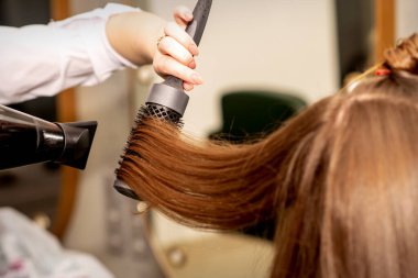 Bir kuaför, uzun kahverengi saçları bir saç kurutma makinesi ve bir güzellik salonunda yuvarlak fırçayla kurutuyor.