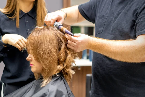 İki kuaför, kuaförde saç maşasıyla uzun kahverengi saç stili yapar.