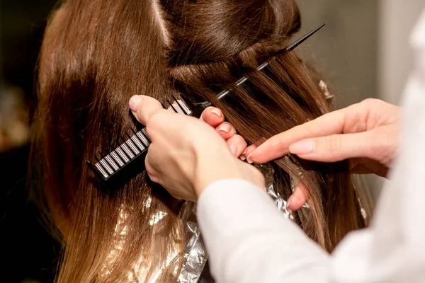 Hairdresser\'s hands prepare brown hair for dyeing with a comb and foil in a beauty salon, close up