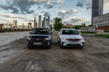 MOSCOW, RUSSIA - 23 Mayıs 2021 Hyundai Creta Birinci nesil (GS / GC) ve Geely Binyue Coolray veya Proton X50 olarak da bilinir. Şehrin arka planında modern bir alt kompakt arazi aracı. Ön taraf görünümü.