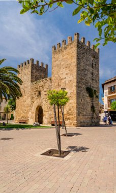 İspanya 'nın her iki tarafında da iki gözetleme kulesi olan tarihi bir kapı. Eski bej duvarlar. Önünde, üzerinde hurma ağaçları ve hurma ağaçları bulunan asfaltlı bir meydan vardır.