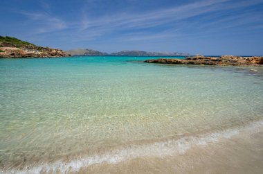 Geniş, terk edilmiş banyo bölmesi kristal berrak, turkuaz su. Körfezi çevreleyen kayalar var. Ufukta daha küçük dağlar görülebilir..