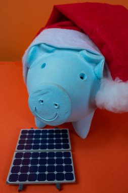 Noel Baba şapkalı domuz kumbarası, fotovoltaik güneş paneli ve turuncu arka planda izole edilmiş lamba..