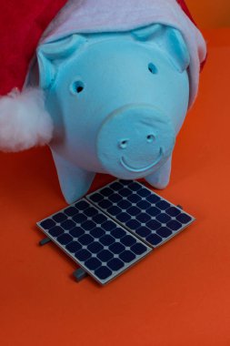 Noel Baba şapkalı domuz kumbarası, fotovoltaik güneş paneli ve turuncu arka planda izole edilmiş lamba..