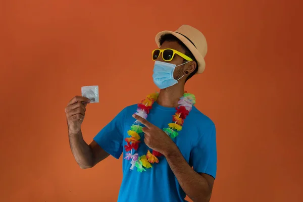 Black man in carnival costume and pandemic mask holding  a condom isolated on orange background. African man in various poses and expressions.