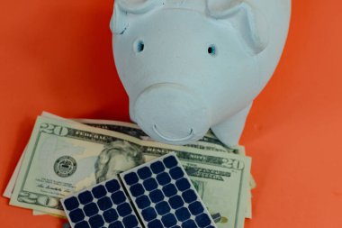 Dolar banknotlu domuz kumbarası ve turuncu arka planda izole edilmiş fotovoltaik güneş paneli. Güneş Enerjisi Kavrama Resmi.