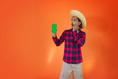 Junina Party Kıyafetli Siyah Adam elinde bir tablet ya da portakal arkaplanda izole edilmiş bir cep telefonu tutuyor. Festa Junina için geleneksel kıyafetler giyen genç adam - Brezilya Haziran Festivali.