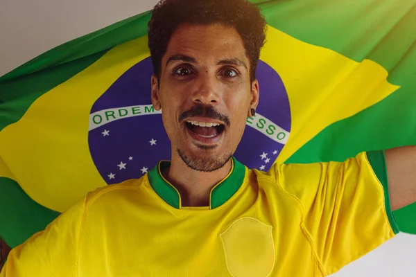 サッカーチームのシャツとブラジルの黒人男性ファンは白に隔離されました カップを祝う旗を持つスポーツファン — ストック写真