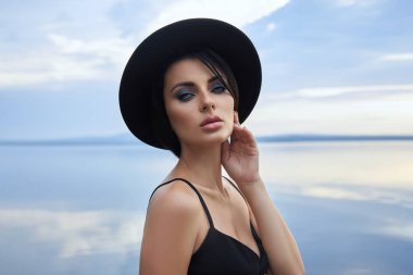 Siyah şapkalı ve siyah elbiseli mükemmel esmer kadın mavi gökyüzüne karşı gölün kenarında poz veriyor. Uzun saçlı kadın ve yüzünde güzel bir makyaj.