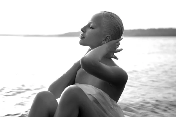 短い散髪とアートヌードセクシーなブロンドは日没時に湖の海岸ビーチに水の中に座っている 濡れた髪と女性の体 保護されたビーチの休日 黒と白 — ストック写真