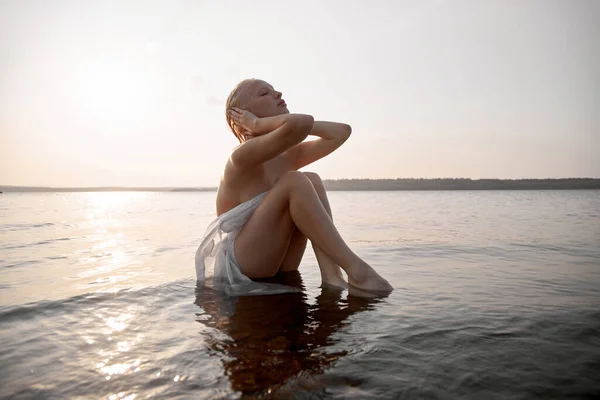 Kısa Saçlı Çıplak Sarışın Gün Batımında Göl Kıyısındaki Sahilde Oturuyor — Stok fotoğraf