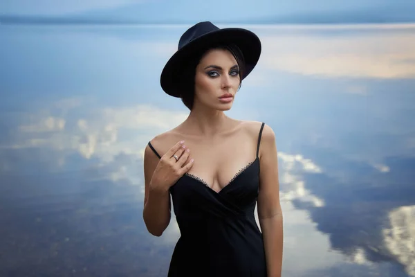 黒の帽子の完璧なブルネットの美しさの女性と黒のドレスは青い空に対して湖の近くにポーズをとります 長いです髪の女性と美しいです化粧上の彼女の顔 — ストック写真