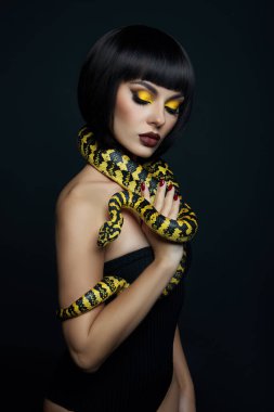 Güzel kadın kısa saç kesimi piton sarısı yılanı boynunda. Bir kızın omuzlarında sarı bir yılan. Güzel sarı göz farı makyajı, koyu kırmızı ruj.