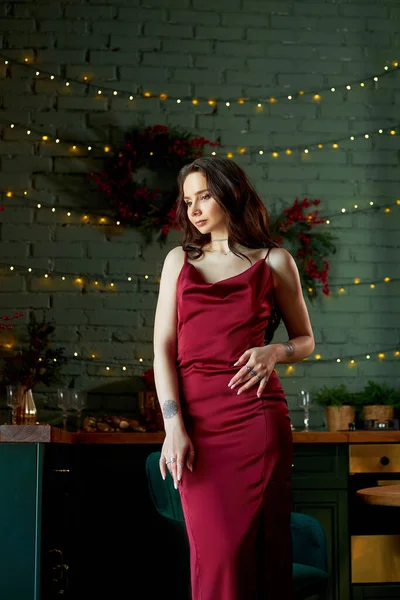 Beautiful woman in red evening light dress at home in the kitchen in the evening. Lights of a garland glow on the wall behind a woman, a beautiful slender body