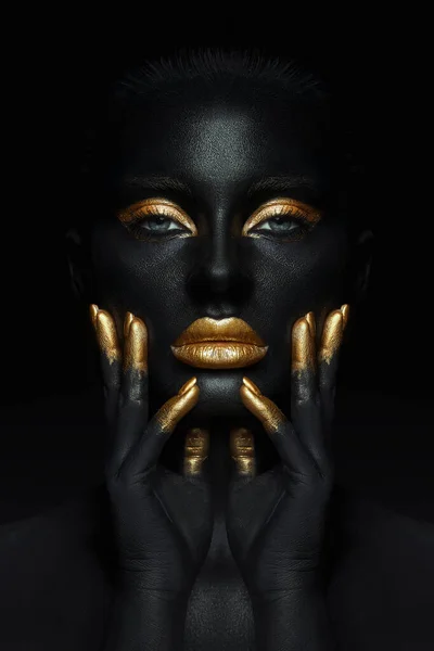 Beauty woman painted in black skin color body art, gold makeup lips eyelids, fingertips nails in gold color paint. Professional gold makeup