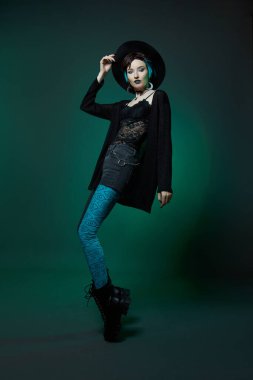 Yuvarlak şapkalı genç cadılar bayramı cadısı ve yeşil siyah arka planda yeşil yuvarlak gözlükler. Boyalı saç, koyu ruj, hilal ay küpeleri.