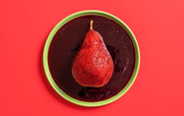 Kırmızı şarap soslu bir tabakta tek bir armut. Armut şarapta kaynatılır, minimalist kırmızı arka planda..