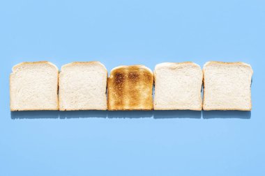 Mavi bir masanın üzerine dizilmiş ekmek dilimleriyle yukarıda. Diğerleri arasında kızarmamış tek bir parça ekmek. Lezzetli tost ekmeği dilimleri mavi arka planda izole edilmiş parlak ışıkta.
