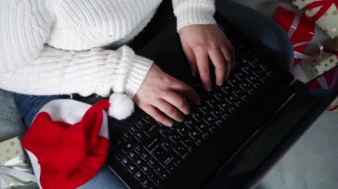 Beyaz kazaklı beyaz kadın, noel hediyeli, Noel Baba şapkalı, Noel ağacı dallı, çalışan, dizüstü bilgisayarda sörf yapan beyaz kadın videosunu kapat.