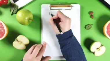 Yakın çekim video kadın elleri notlarda yemek diyet planı yazar, sağlıklı meyve, sebze, tablet, not defteri ve aksesuarlar parlak yeşil masa arka planında
