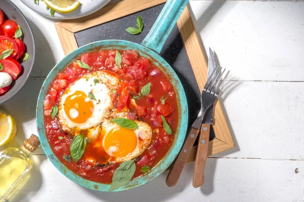 Traditional greek food, Shakshouka. eggs poached in frying pan with tomatoes, olive oil, peppers, onion, garlic, Mediterranean cuisine. Keto, fodmap low carb diet recipe, white wooden background