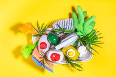 Yaz temizliği konsepti. Plastik sepet, farklı deterjanlar ve temizlik malzemeleri şişeleri, parlak sarı arka planda palmiye yapraklı temizlik için aksesuarlar.