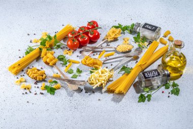 Beyaz masa arkasında çeşitli kurutulmuş spagetti, makarna ve kaşıklarla hazırlanmış makarna. Farklı şekillerde çiğ İtalyan makarnası, otlar, zeytinyağı, domatesler, baharatlar.