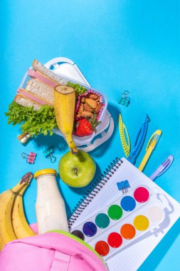 Sağlıklı okul beslenme çantası: sandviç, sebze, meyve, fındık ve yoğurt, içinde okul çocukları malzemeleri, aksesuarlar ve yüksek renkli mavi arka plan fotokopi alanı üzerine sırt çantası